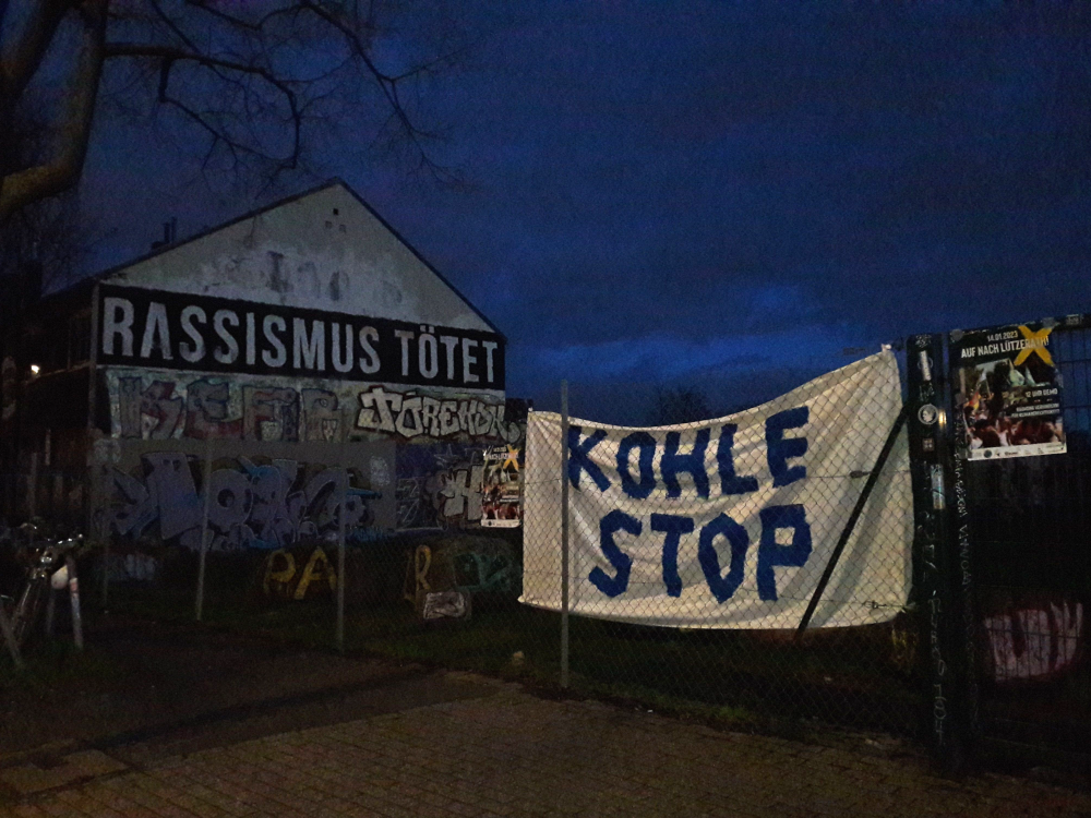 Banner "Kohle Stop" am Zaun der Brache neben dem AZ Köln, im Hintergrund die Westfassade mit dem Schriftzug "Rassismus tötet"