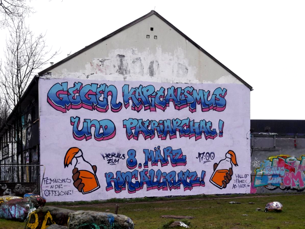 Graffiti-Wandbild an der Seitenfassade des Autonomen Zentrums Köln: "Gegen Kapitalismus und Patriarchat! Heraus zum 8. März, 17 Uhr, Roncalliplatz. Feminismus in die Offensive!". Daneben jeweils eine gesprayte Hand, die eine Flasche mit orangener Flüssigkeit und Tuchverschluss darsellt.
